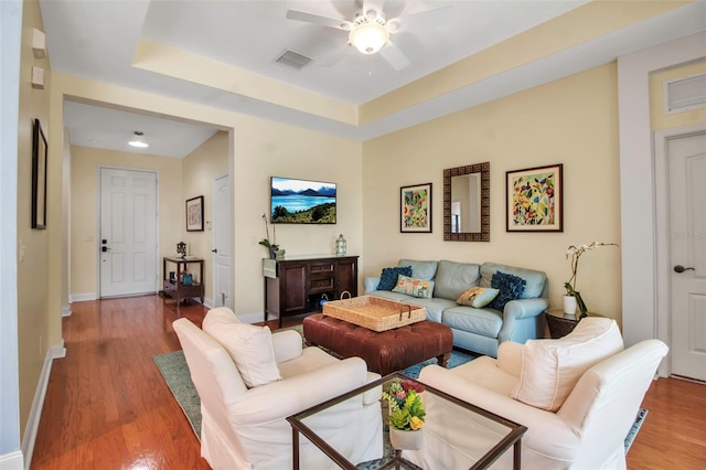 living area with ceiling fan, wood finished floors, baseboards, and a tray ceiling