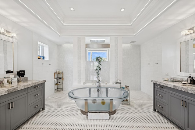 bathroom featuring tile walls, a soaking tub, a tray ceiling, two vanities, and crown molding