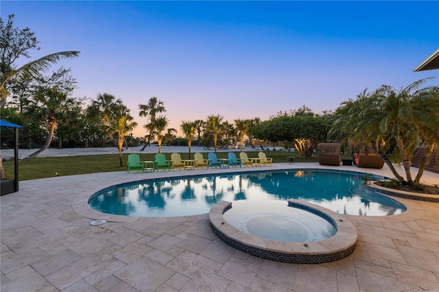 pool at dusk featuring a patio area, a pool with connected hot tub, and a yard