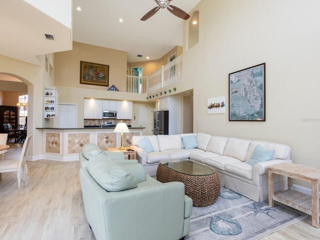 living room with arched walkways, ceiling fan, light wood-style flooring, a towering ceiling, and recessed lighting