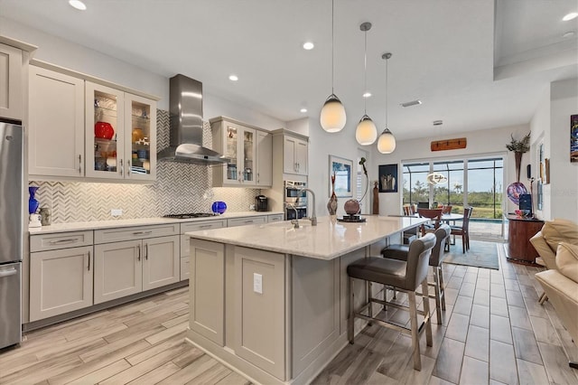 kitchen featuring wall chimney range hood, decorative backsplash, open floor plan, an island with sink, and light wood finished floors
