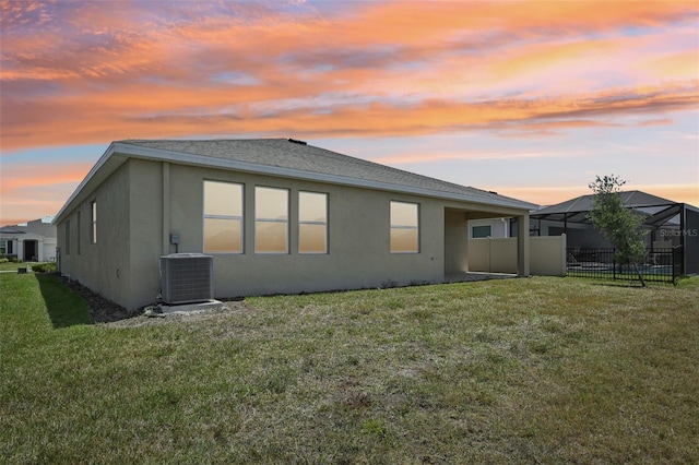 back of property at dusk with stucco siding, central AC unit, a lawn, and a shingled roof