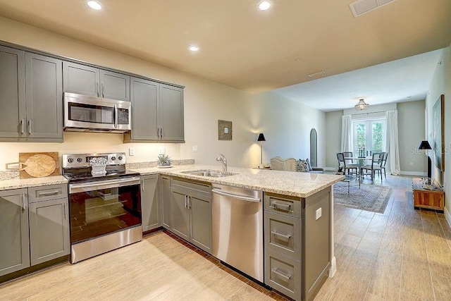 kitchen featuring appliances with stainless steel finishes, a sink, gray cabinetry, a peninsula, and light stone counters