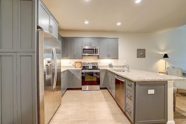 kitchen with stainless steel appliances, gray cabinets, a peninsula, a sink, and light stone counters