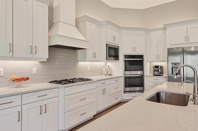 kitchen featuring stainless steel appliances, a sink, premium range hood, crown molding, and white cabinetry