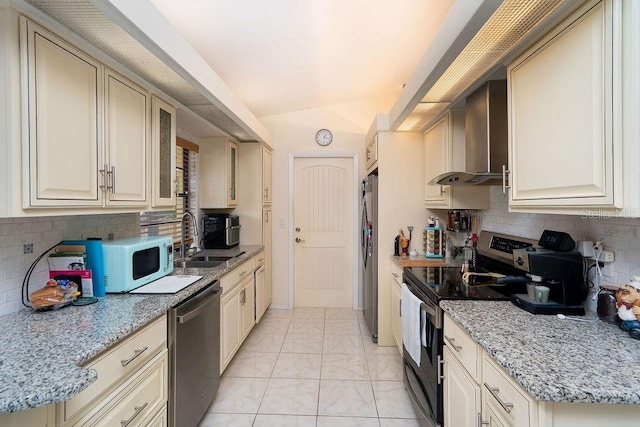 kitchen with stainless steel appliances, a sink, wall chimney exhaust hood, cream cabinetry, and vaulted ceiling