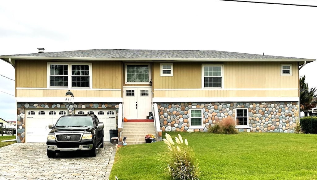 5111 Bay State Rd, Palmetto, FL 34221 - view of front of home with decorative driveway, a front lawn, stone siding, an attached garage, and a shingled roof