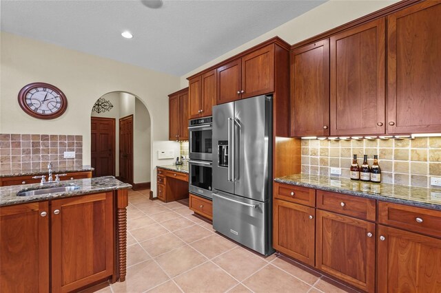 7025 Saddle Creek Cir, Sarasota, FL 34241 - kitchen with stainless steel appliances, arched walkways, decorative backsplash, dark stone countertops, and brown cabinetry