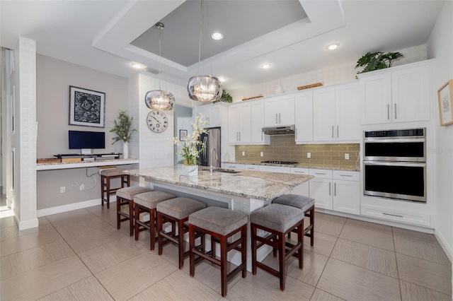 826 Honeyflower Loop, Bradenton, FL 34212 - kitchen with under cabinet range hood, stainless steel appliances, a raised ceiling, decorative backsplash, and a breakfast bar area
