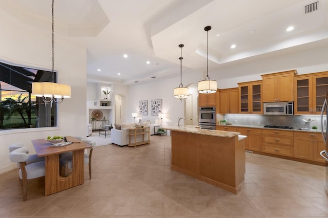 7144 Ashland Gln, Lakewood Ranch, FL 34202 - kitchen with appliances with stainless steel finishes, a raised ceiling, tasteful backsplash, light stone counters, and a center island with sink