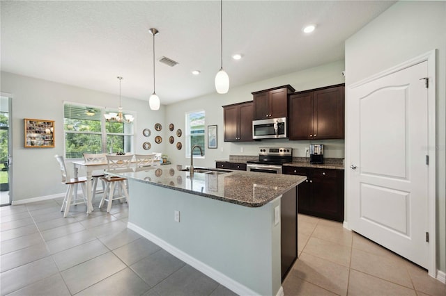 13804 Woodbridge Ter, Lakewood Ranch, FL 34211 - kitchen with stainless steel appliances, dark stone counters, dark brown cabinets, an island with sink, and light tile patterned floors