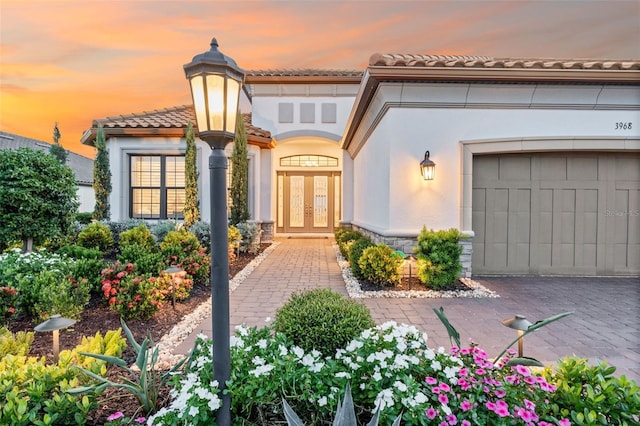 3968 Santa Caterina Blvd, Bradenton, FL 34211 - exterior entry at dusk with a tiled roof, stucco siding, french doors, an attached garage, and stone siding