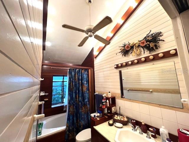 full bathroom featuring toilet, ceiling fan, vanity, wooden walls, and shower / tub combo with curtain