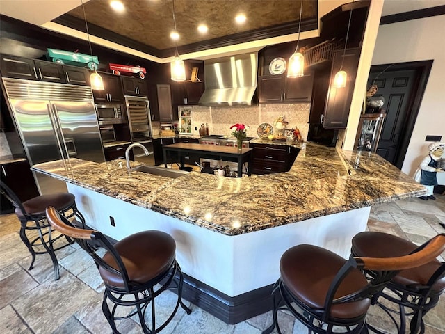 kitchen with wall chimney exhaust hood, appliances with stainless steel finishes, decorative light fixtures, a breakfast bar, and stone tile floors