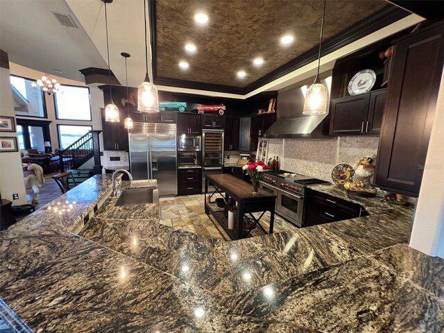 kitchen featuring backsplash, decorative light fixtures, wall chimney exhaust hood, sink, and built in appliances