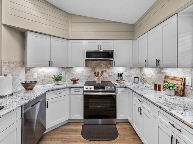 kitchen with white cabinets, stainless steel appliances, vaulted ceiling, light stone counters, and light hardwood / wood-style flooring