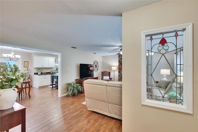 living area with visible vents, a textured ceiling, light wood-type flooring, baseboards, and ceiling fan with notable chandelier