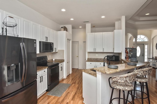 kitchen with black appliances, stone counters, a kitchen bar, white cabinetry, and light wood-type flooring