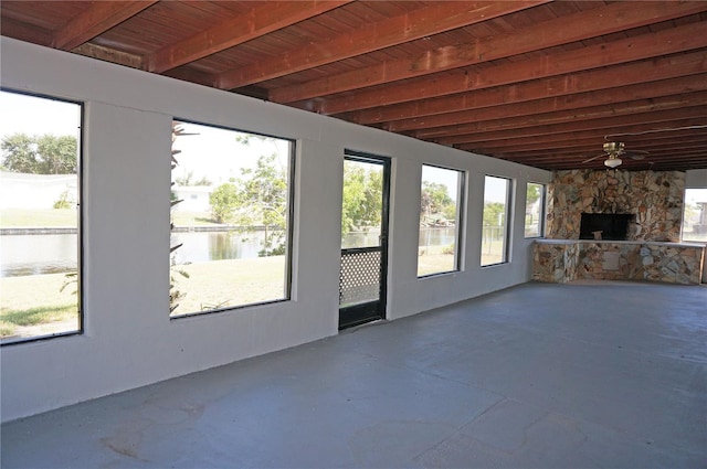 unfurnished sunroom with ceiling fan, wood ceiling, a wealth of natural light, a stone fireplace, and a water view