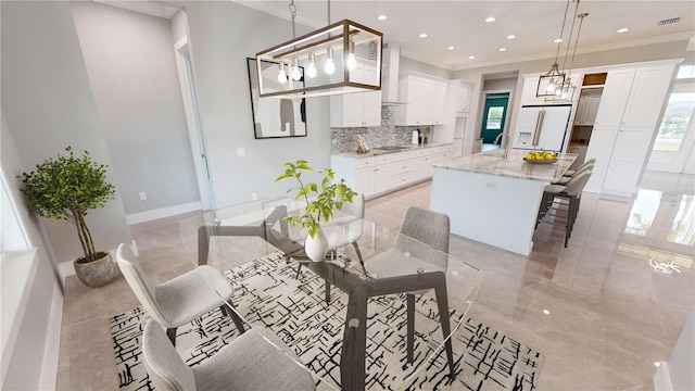 kitchen with wall chimney exhaust hood, white refrigerator with ice dispenser, hanging light fixtures, light stone counters, and a center island with sink