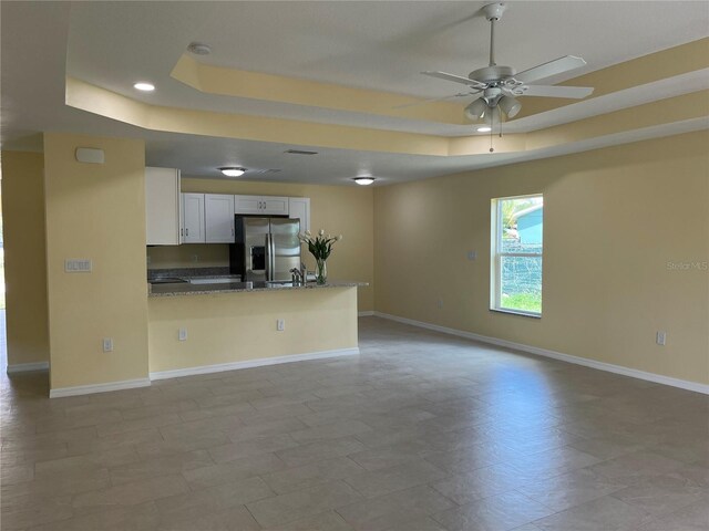 kitchen with white cabinetry, stainless steel fridge with ice dispenser, a tray ceiling, kitchen peninsula, and dark stone counters