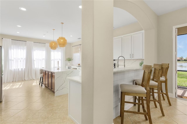 kitchen with pendant lighting, a kitchen breakfast bar, light tile flooring, light stone countertops, and white cabinetry