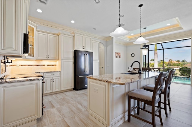 kitchen with an island with sink, a tray ceiling, high end black refrigerator, hanging light fixtures, and light stone counters