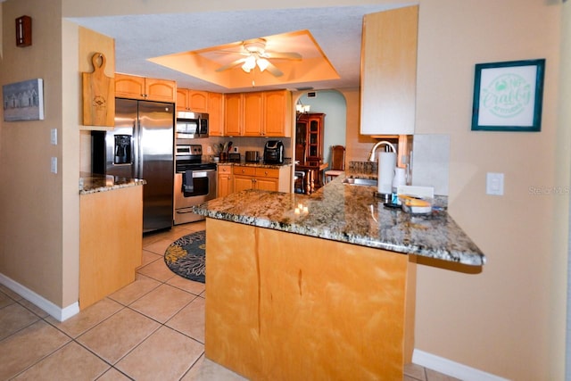 kitchen with a tray ceiling, ceiling fan, appliances with stainless steel finishes, sink, and dark stone counters