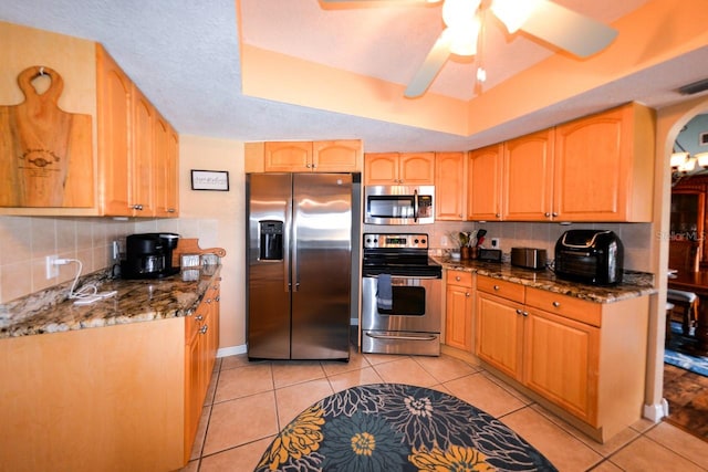 kitchen with ceiling fan, dark stone counters, appliances with stainless steel finishes, light tile flooring, and tasteful backsplash