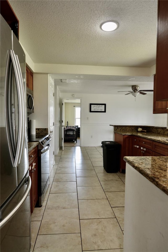 kitchen featuring appliances with stainless steel finishes, light tile floors, dark stone countertops, ceiling fan, and a textured ceiling