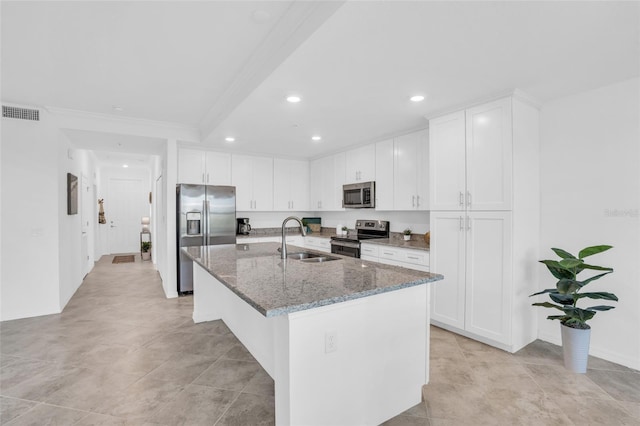 kitchen with dark stone counters, a kitchen island with sink, stainless steel appliances, white cabinetry, and a sink