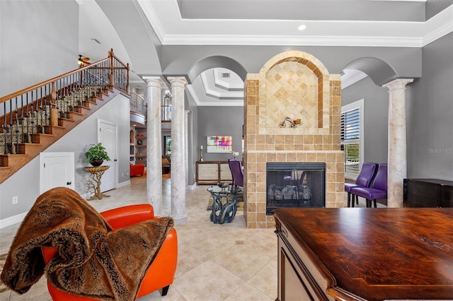 living room featuring light tile floors, a tray ceiling, a tiled fireplace, decorative columns, and crown molding