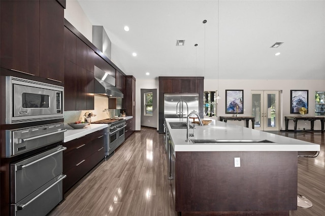 kitchen featuring built in appliances, decorative light fixtures, wood-type flooring, tasteful backsplash, and a kitchen island with sink