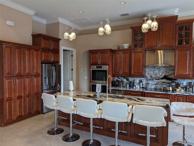 kitchen with wall chimney range hood, backsplash, an island with sink, stainless steel appliances, and pendant lighting