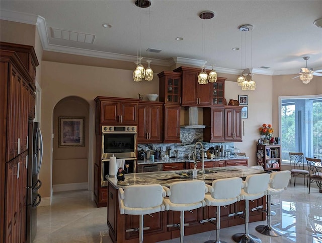 kitchen featuring ceiling fan with notable chandelier, pendant lighting, wall chimney exhaust hood, a center island with sink, and a breakfast bar