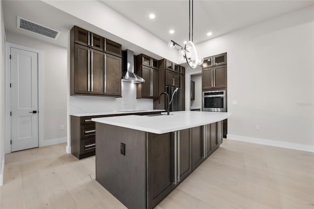 kitchen featuring pendant lighting, wall chimney range hood, dark brown cabinets, an island with sink, and stainless steel oven