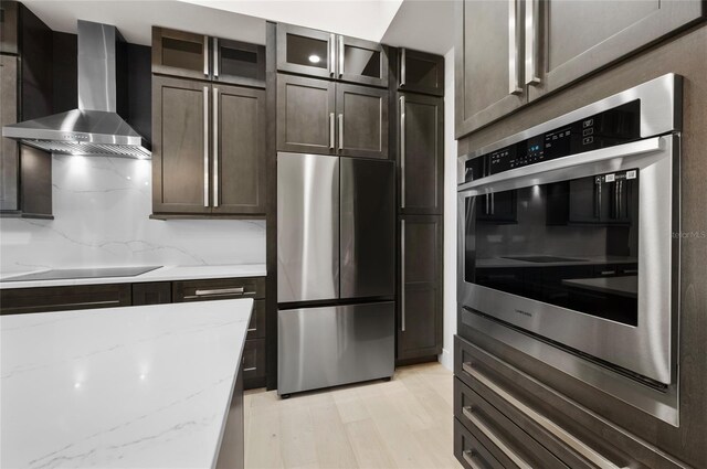 kitchen featuring dark brown cabinetry, light stone counters, appliances with stainless steel finishes, wall chimney range hood, and backsplash