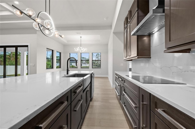 kitchen with pendant lighting, black electric stovetop, a raised ceiling, light stone countertops, and wall chimney range hood