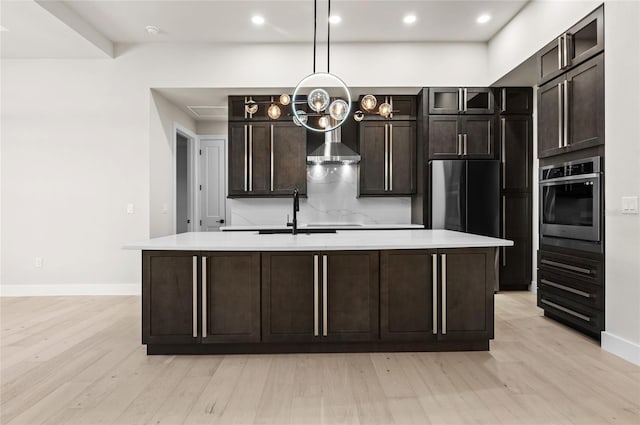 kitchen with dark brown cabinetry, light wood-type flooring, stainless steel appliances, a kitchen island with sink, and wall chimney range hood