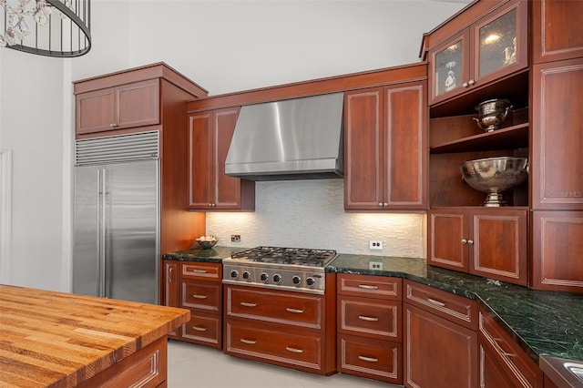 kitchen with dark stone countertops, appliances with stainless steel finishes, wall chimney exhaust hood, backsplash, and light tile flooring