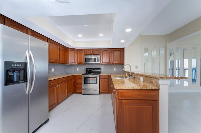 kitchen with kitchen peninsula, appliances with stainless steel finishes, light stone counters, a raised ceiling, and sink