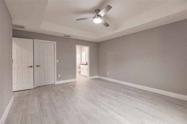 unfurnished bedroom featuring ensuite bathroom, a raised ceiling, light hardwood / wood-style flooring, ceiling fan, and a closet