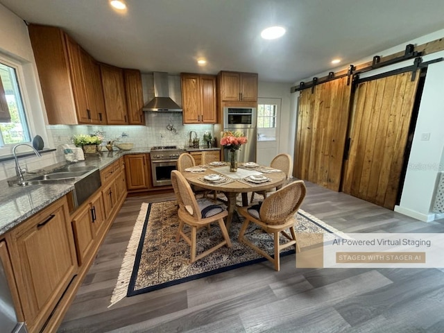 kitchen featuring wall chimney range hood, sink, tasteful backsplash, light stone counters, and a barn door