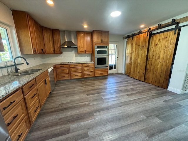 kitchen with sink, appliances with stainless steel finishes, light stone counters, a barn door, and wall chimney exhaust hood