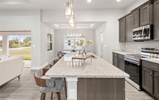 kitchen with an inviting chandelier, a raised ceiling, an island with sink, appliances with stainless steel finishes, and light stone counters