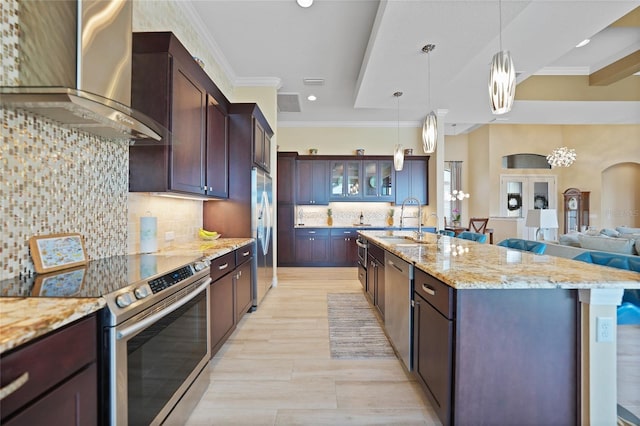 kitchen featuring dark brown cabinetry, wall chimney exhaust hood, hanging light fixtures, stainless steel appliances, and a kitchen island with sink