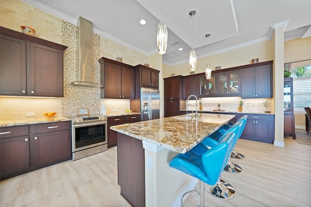 kitchen with sink, wall chimney exhaust hood, stainless steel appliances, light stone counters, and a center island with sink