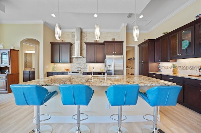 kitchen featuring hanging light fixtures, stainless steel appliances, wall chimney range hood, and a center island with sink