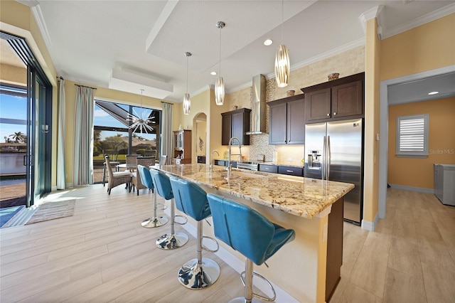 kitchen with wall chimney range hood, hanging light fixtures, stainless steel fridge, an island with sink, and dark brown cabinetry