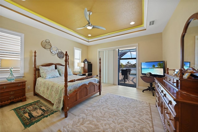 bedroom featuring access to outside, light hardwood / wood-style flooring, ceiling fan, a textured ceiling, and a tray ceiling